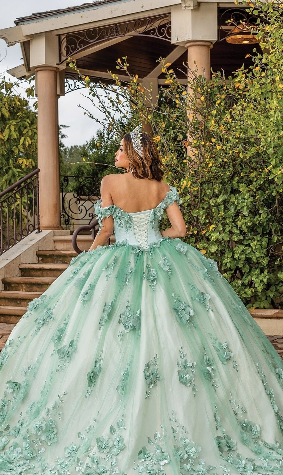 A woman stands on a brick path in front of a pavilion, facing away, wearing the Quinceanera Dress 1818 By Dancing Queen—a light green, off-the-shoulder ball gown with floral appliqués and an A-line silhouette.